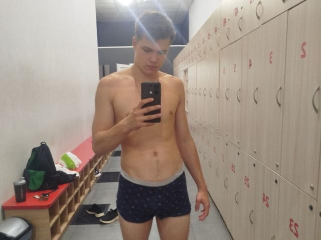 LucasLong stands in a locker room, shirtless, wearing dark blue boxer shorts with a pattern, holding a phone, looking at it, with a slight frown, reflecting in a mirror.