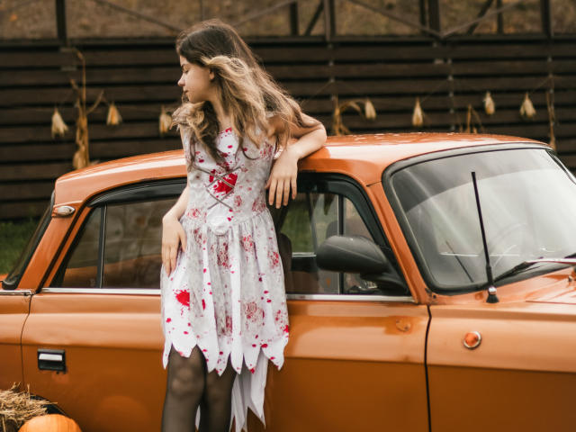 TinaRedly leans against an orange vintage car, wearing a white floral dress with red roses and black tights. Her wavy, brown-to-blonde ombre hair cascades over her shoulders. She rests her right arm on the car's roof, her left hand on her hip. The car's glossy paint contrasts with the dress's delicate fabric. String lights hang in the background. Her relaxed, sideways glance adds a casual vibe. The image has a natural, slightly overcast light.