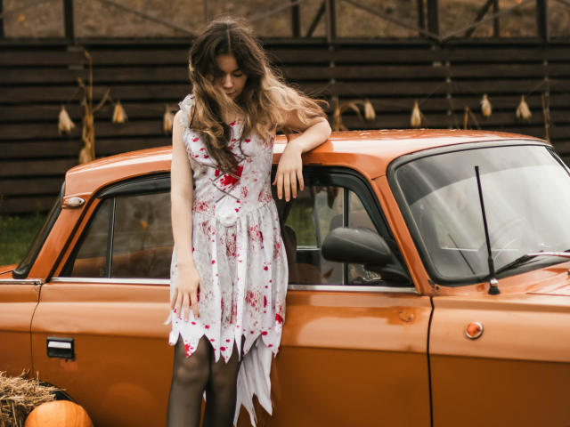 TinaRedly stands beside a rusty orange vintage car, leaning on the roof with her right arm. She has wavy, brown hair and wears a white dress with red floral patterns and black tights. Her left hand rests on the car's door. The car's textured paint and weathered surface contrast with her delicate, floral dress. The image uses natural light, highlighting her relaxed posture and the car's classic, worn-out style. The background features a dark wooden fence with hanging dried plants.