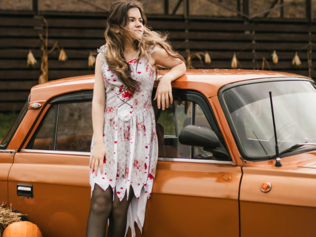 TinaRedly stands leaning on an orange vintage car. She has long, wavy brown hair and wears a white floral dress with red hearts, black stockings, and a white bow. Her right arm rests on the car's roof, and her left hand touches the window. A small pumpkin is on the ground. The background features a dark wooden fence with dried corn cobs. The image has a warm, autumnal feel.