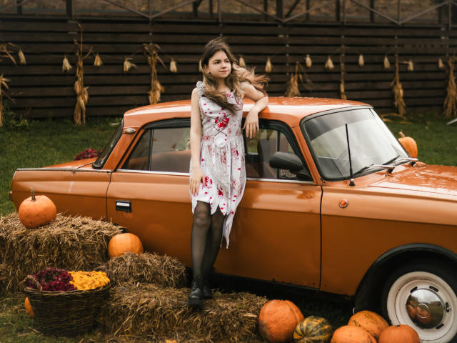 A photo of TinaRedly leaning against an orange vintage car. She's wearing a white floral dress with red stains, black tights, and black boots. Her long brown hair is slightly windswept. Surrounding the car are hay bales, baskets of colorful squash, and several pumpkins. The car's shiny orange paint contrasts with the muted autumnal colors. TinaRedly's relaxed posture and direct gaze are key elements. The image uses natural light and has a rustic, autumnal vibe. The overall composition is balanced, with TinaRedly as the focal point.