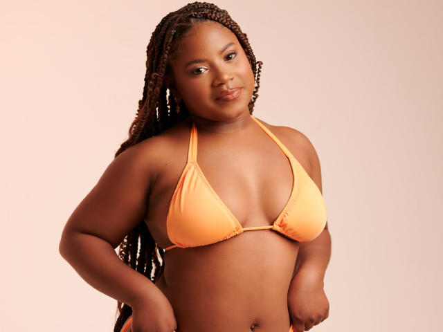 PrettyChocolate met donkerbruine huid, lang gevlochten haar, oranje bikini top, waarbij haar kleine borsten zichtbaar zijn, en een zelfverzekerde glimlach, handen op de heupen, staand voor een effen achtergrond..