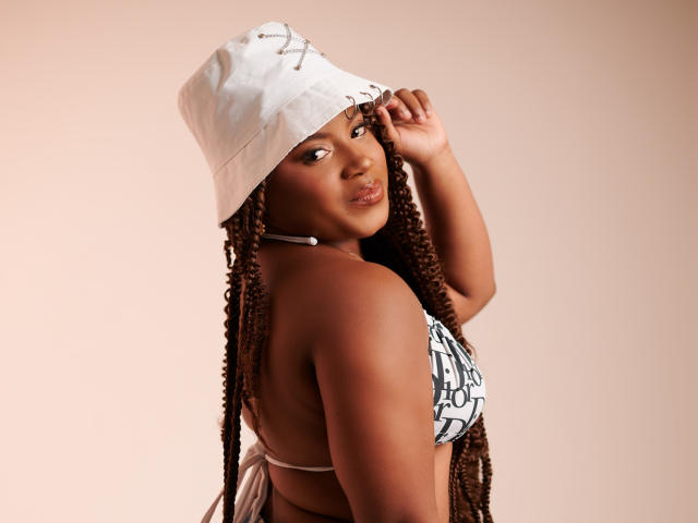 PrettyChocolate stands in a three-quarter profile, facing right. She has dark brown, braided hair extending down her back, wearing a white, slightly worn bucket hat. She wears a black-and-white strapless top, revealing her shoulders and upper back. Her right hand is gently holding the hat's brim. She has a smooth, dark brown complexion and a neutral, slightly pensive expression. The soft, beige background is plain and untextured. The image has a clean, modern style with a focus on PrettyChocolate's confident, relaxed posture.