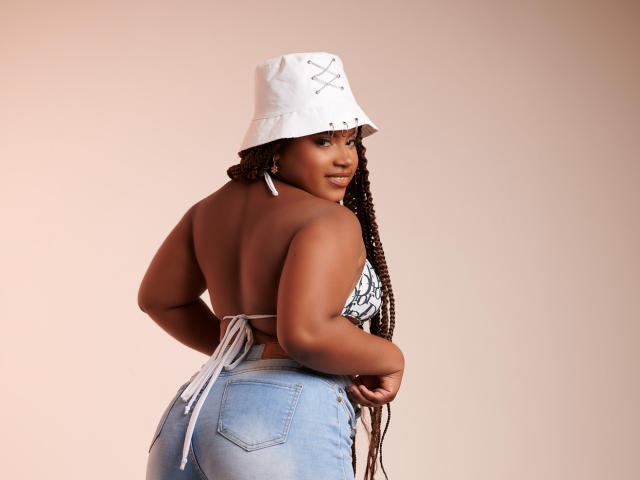 PrettyChocolate, a dark-skinned woman with long braided hair, stands with her back to the camera. She wears a white bucket hat with black stitching and a black-and-white strapless top that reveals her back. Her blue jeans are high-waisted with a white tie at the back. She looks over her shoulder with a slight smile. The image focuses on her curvy, muscular build and confident posture. The lighting is even, highlighting her smooth, dark skin and the texture of her hair.