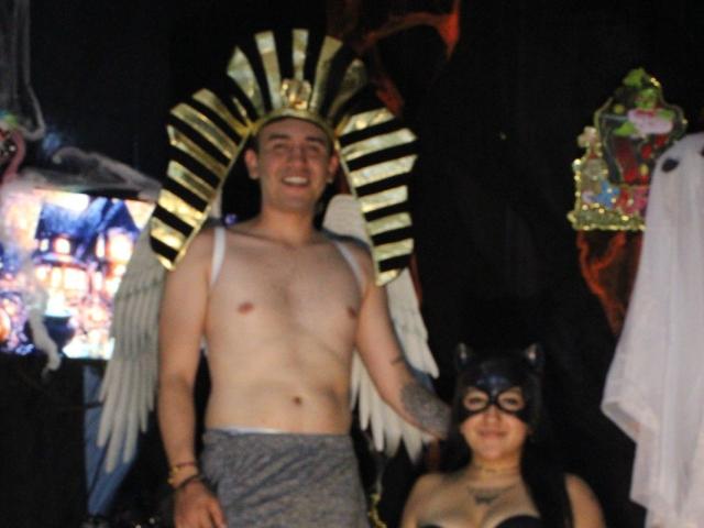 TemptationDual con ali d'angelo e copricapo dorato, senza camicia, con un drappo grigio, sorridente. TemptationDual con maschera da gatto nera, capelli neri, sorridente, in piedi. Entrambe in un ambiente scuro e poco illuminato..