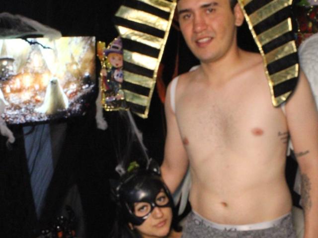 Tentazione sensuale: uomo senza camicia con copricapo egizio dorato, pantaloncini grigi, tatuaggio sul braccio destro e una maschera nera accanto a lui. Persona mascherata in costume nero con orecchie da gatto..