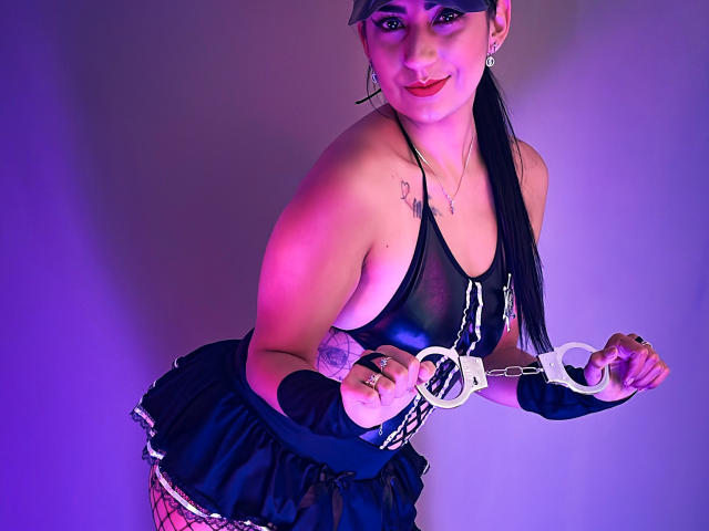 PaullieEvans in black lingerie, handcuffs, and a headmask, showing tattoos, with a black skirt, fishnet, and a pink-purple light. She's smiling, with long black hair.