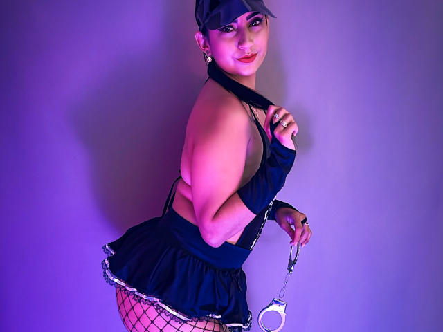 Paullie Evans, con un cuerpo atractivo y curvilíneo, lleva puesto un gorro negro, un top de tirantes, una minifalda, medias de red y esposas, y sonríe a la cámara. Iluminación morada y rosa..