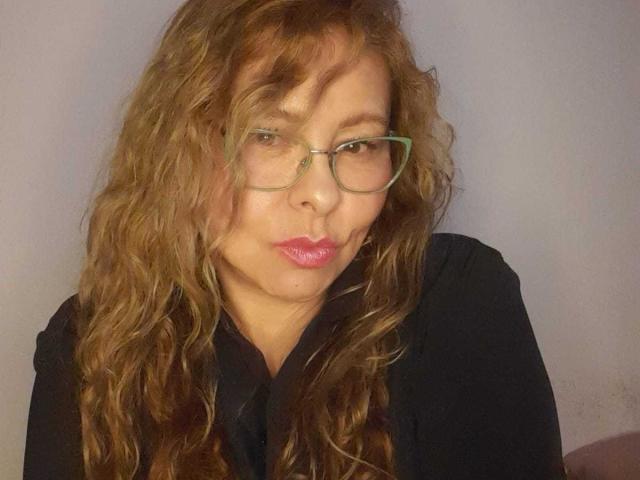 MiaAmoretti with wavy brown hair, green glasses, and pink lipstick, pouting her glossy lips, wearing a black shirt. Her tits are small, and she's looking cute.