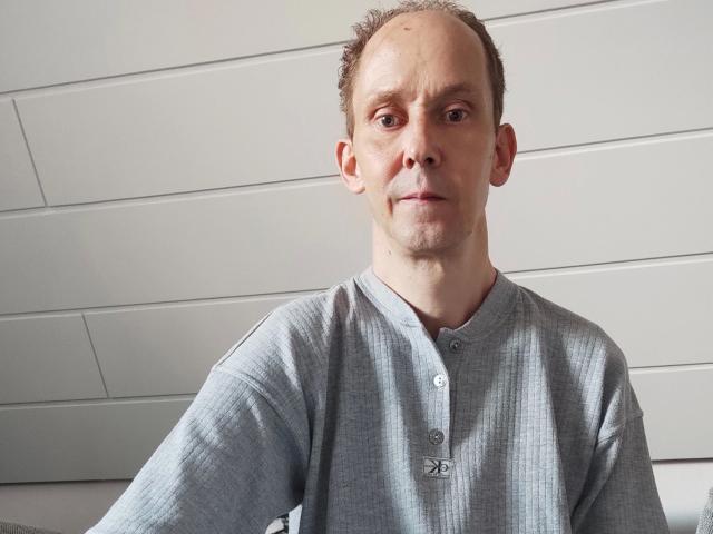 Sveniboy1-hot, bald with short brown hair, wearing a gray button-up shirt, has a serious expression, and is standing against a white wall. Slightly wrinkled shirt.