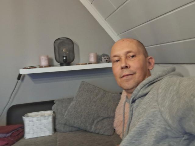 Sveniboy1-hot, bald with light skin, wearing a gray hoodie, smiling, sitting on a gray couch with a white basket and candles on a shelf behind him.