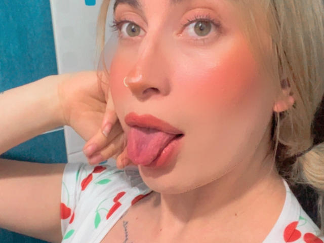 SammyViolet with blonde hair, green eyes, and a nose ring, sticking out her pink tongue, wearing a white cherry print shirt, looking at the camera.