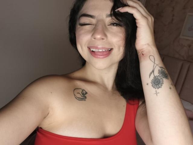 AnntonellaWatson with black hair, winking, smiling, red top, showing tattoos on right arm and left chest, right hand in hair, small cut on right arm.