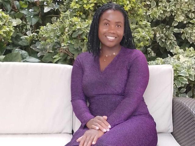 CreamPieQueen sits on a white cushioned sofa. She has dark skin, and her hair is in tight, small twists. She's wearing a purple, long-sleeve, V-neck dress. Her hands are clasped together in her lap. She's smiling with white teeth. The background is green leafy bushes. The texture of her dress is slightly shiny. She's centered in the image. The overall composition is simple and focused on her.