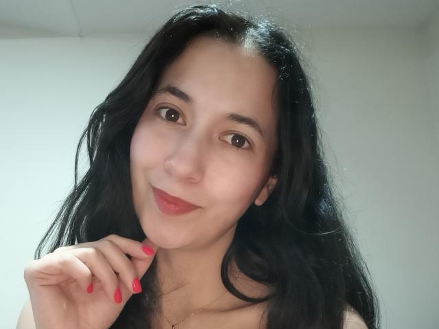 AnneStone with long black hair, brown eyes, and red lipstick, smiling slightly, holding her chin with pink-manicured fingers, looking cute and sexy.