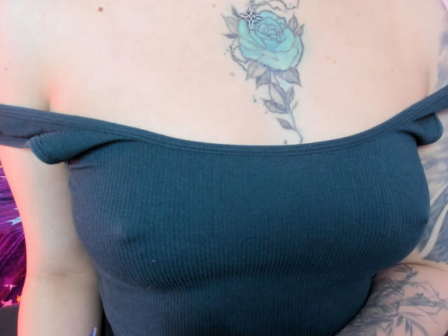 KayleeM's tits in a ribbed, dark green off-shoulder top, showing a blue rose tattoo on her chest. Her arm has a tattoo, too.
