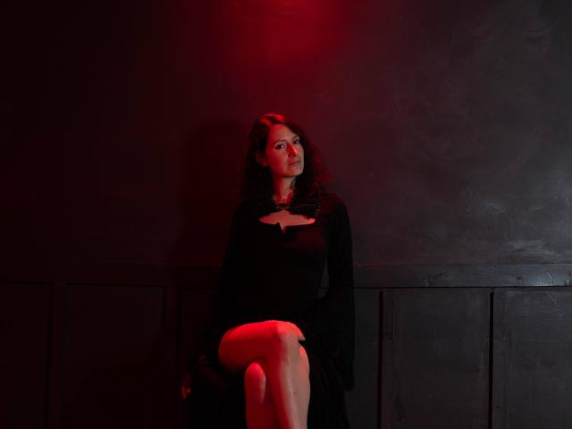 MistressLucia, avec ses cheveux noirs bouclés, portant une robe noire avec une découpe, assise les jambes croisées, une lumière rouge projetant des ombres, regardant légèrement vers le haut, dans un décor sombre et mélancolique..