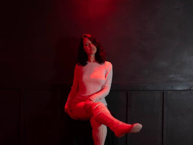 MistressLucia sits on a black stool, legs crossed, in a dimly lit room with red lighting. She wears a light pink, long-sleeve dress with white, quilted thigh-high boots. Her dark curly hair falls to the sides, and she looks slightly to the right. The red light casts a subtle glow on her face and dress, highlighting her relaxed, yet poised, posture. The texture of the dress contrasts with the quilted boots and the smooth, dark wall behind her. The composition emphasizes her form and the interplay of light and shadow.