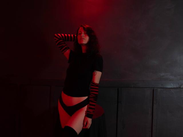 MistressLucia, avec ses cheveux bouclés, son haut noir, ses manchettes rayées, sa culotte noire et ses bas montants, se tient devant un mur sombre, une lumière rouge projetant une ambiance mélancolique, avec une main sur sa tête..