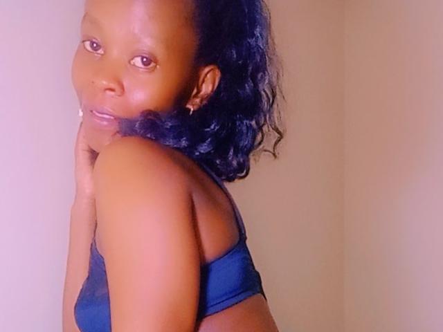 PelgyCarter with dark brown skin, curly black hair, small hoop earrings, and wearing a blue bralette, looking over her shoulder with a subtle smile.