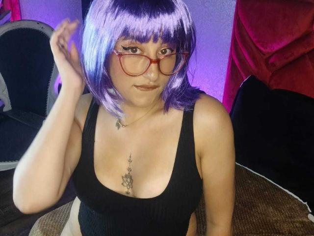 JudyCampbells with purple bob, red glasses, black tank top, showing tits with a tattoo between, hand in hair, sitting, red velvet background, soft purple lighting.