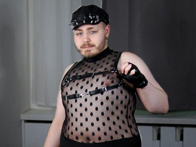 FabianNoLimits with a black leather cap, black fingerless gloves, and a sheer polka-dot top showing his bare chest and nipples, standing in a gray room.