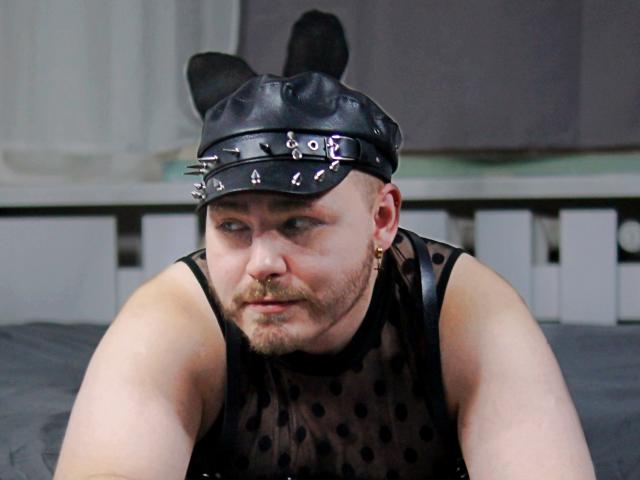 FabianNoLimits with a bald head, black leather cap with studs, black mesh polka-dot top, and black bunny ears, looking down with a serious expression, sitting on a bed.