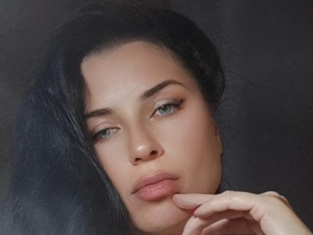 RosannaLora's face with dark, wavy hair, blue eyes, and pink lips, lightly touching her chin with a subtle, sexy smile, soft skin, and a slightly flushed nose.