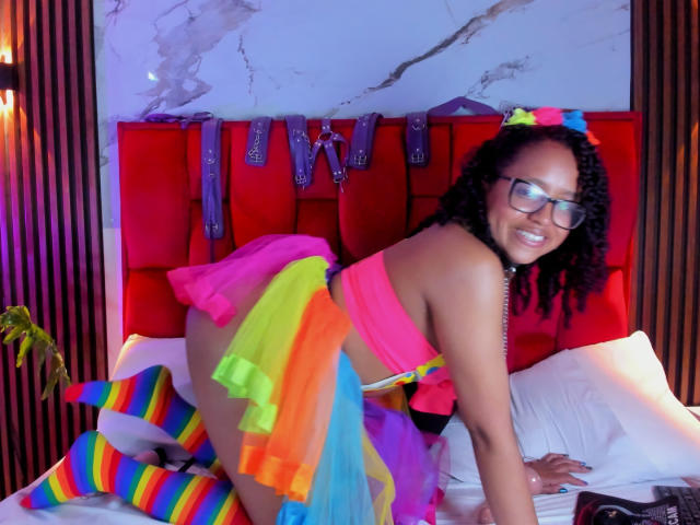 ArtemisaS with curly black hair, glasses, smiling, wearing a pink top, rainbow tutu, and rainbow-striped socks, kneeling on a bed with a red headboard. Her ass is raised.