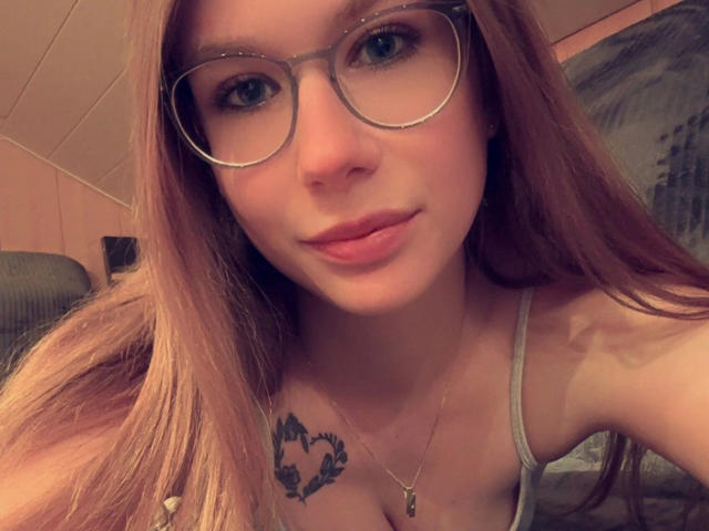 Soleea-hot with long brown hair, glasses, and pink lips, showing a small heart tattoo above her tits. She's wearing a grey top and a necklace.