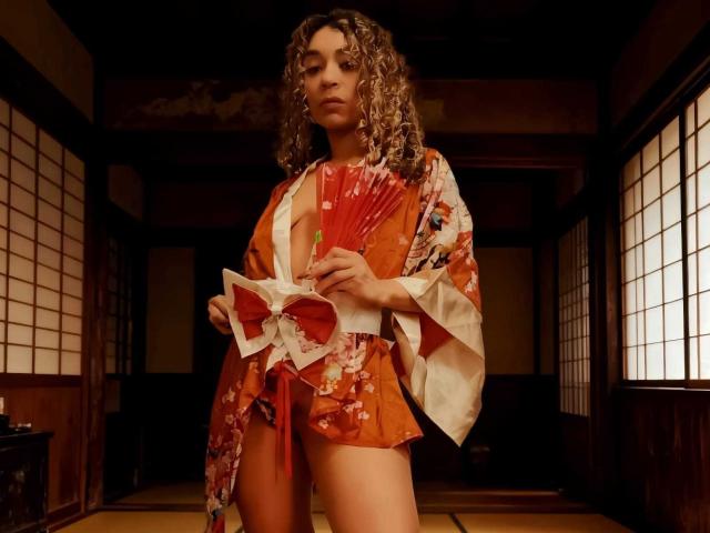 KylieJen with curly brown hair, wearing an orange floral kimono, exposing her small tits, holding a red fan, standing in a dimly lit room with wooden shoji screens.