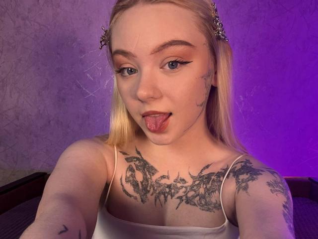EnchantingEyes with blonde hair, stick-on butterfly hair clips, tattoos on chest and arms, sticking out tongue, wearing white spaghetti strap top, under purple light.