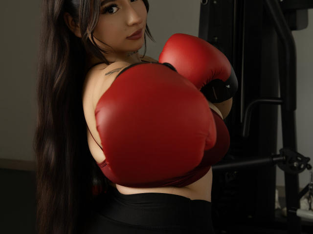 MiaSuarez with long dark hair, red boxing gloves, and black workout pants, showing her tits, standing in a gym, looking at the camera with a serious expression.