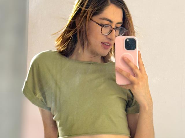 RousseBlossom, a young woman with fair skin and brown hair, takes a mirror selfie. She wears round glasses and a light green, short-sleeved crop top. She holds a pink phone with a white ring in her right hand. Her hair is slightly tousled, and she looks down at the phone with a subtle smile. The image is sharply focused on her upper body and face. Natural light highlights her features.