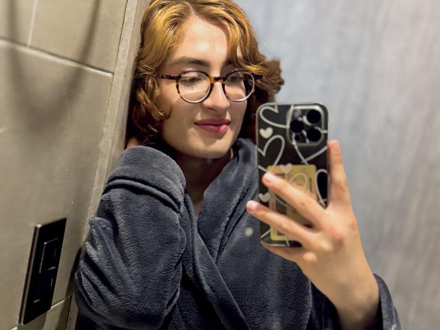 RousseBlossom takes a mirror selfie. They have wavy, shoulder-length red hair and wear round glasses. They're dressed in a dark blue, textured bathrobe. Their right hand holds a phone with a black and white patterned case. The phone case has a gold accent. RousseBlossom has a slight, confident smile. The photo is sharply focused, with a soft, even light. The background is blurred, emphasizing RousseBlossom's face and phone.