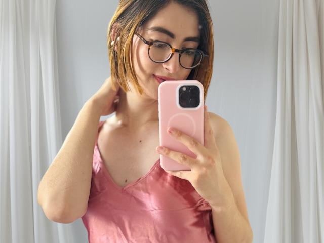 RousseBlossom with short brown hair, glasses, pink satin top, holding pink phone, taking mirror selfie, smiling, hand touching neck.