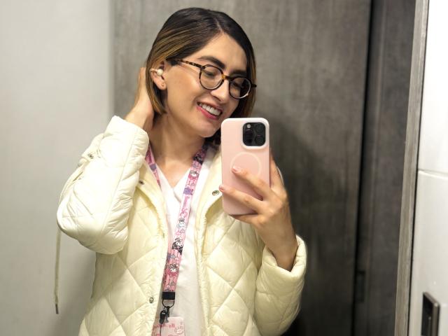 RousseBlossom con gafas, sonriendo, sosteniendo un teléfono rosa, vistiendo una chaqueta blanca acolchada, un lanyard floral rosa, cabello castaño corto, mano en la cabeza, selfie en el espejo, dientes visibles..