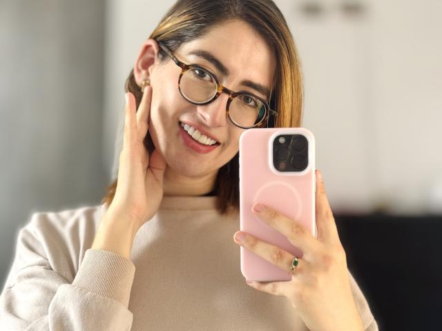 RousseBlossom with glasses, smiling, holding pink phone, pink ring, beige sweater, hands on cheeks, brown hair, white background, soft light, casual selfie, cute, attractive.
