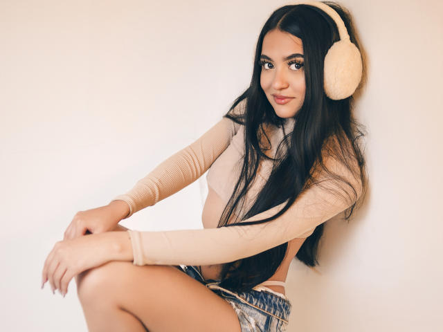 AmySaens, con cabello negro largo, usando auriculares beige, una blusa beige de manga larga y pantalones cortos vaqueros, sentada con una ligera sonrisa, mostrando sus pequeños senos y su lindo trasero..