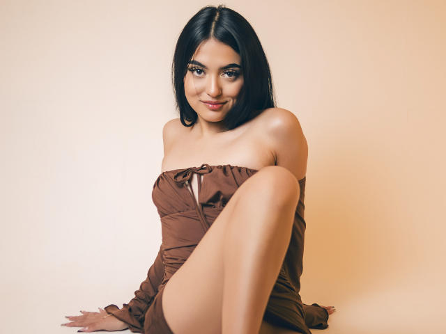 AmySaens has shoulder-length black hair and a fair skin tone. She's sitting with one leg bent, wearing an off-shoulder brown dress that's slightly wrinkled. Her dark brown eyes are slightly squinted, and she's smiling softly. Her right arm rests on her bent knee, while her left arm is on the floor. The dress has a small bow at the center. The image has a warm, beige background. She has a slim, attractive build. The lighting is even, highlighting her smooth skin and subtle makeup. The composition focuses on her upper body and face.