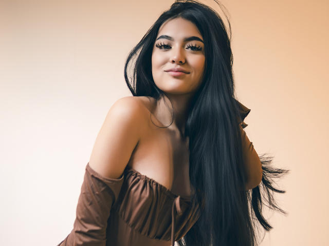 AmySaens has long, straight black hair that flows over her right shoulder. She's wearing an off-shoulder, brown top that reveals her shoulders and upper chest. Her eyebrows are thick and well-defined, and she has long, curled eyelashes. Her skin tone is warm and smooth. She has a slight, confident smile. The image is softly lit with a warm, beige background. The composition focuses on her upper body and face, with her hair taking up a significant portion of the frame.