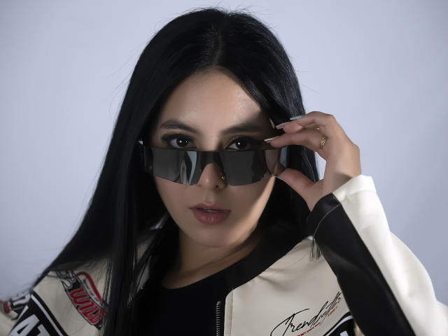 EimmyPark with long black hair, wearing a white and black jacket with red and black designs, black shirt, and black sunglasses, looking at camera, hand adjusting sunglasses.