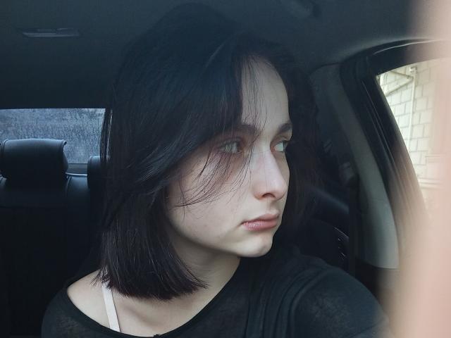 Elissfan with pale skin, black shoulder-length hair, and green eyes, wearing a black shirt, looking out of a car window with a serious expression, slight wind in her hair.