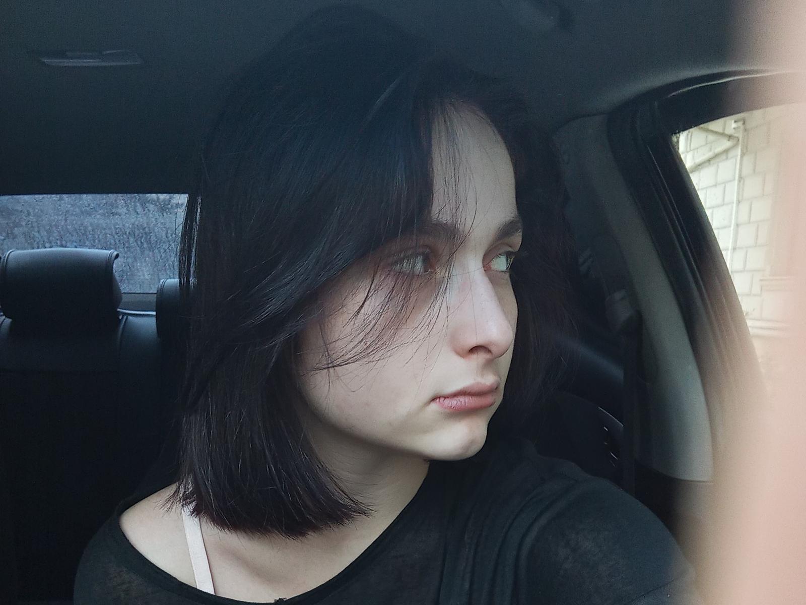 Elissfan with pale skin, black shoulder-length hair, and green eyes, wearing a black shirt, looking out of a car window with a serious expression, slight wind in her hair.
