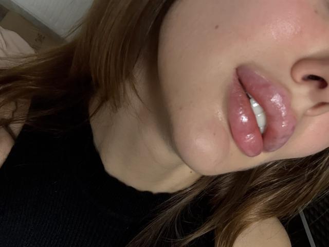 SensualMila's glossy, pink, plump lips are slightly parted, showing a hint of white teeth. Her fair skin has a slight sheen, and brown hair is partially visible.