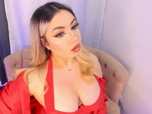 MelissaLust with blonde hair, red lipstick, and heavy eyeliner, wearing a red dress that shows her large boobs. She's sitting in a beige chair, looking up.
