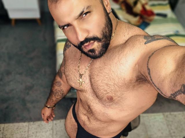 BardoCkroy69 is a hairy, bearded, tattooed, muscular dude wearing a black thong, gold cross necklace, and a bracelet, taking a selfie with a serious, sexy expression.