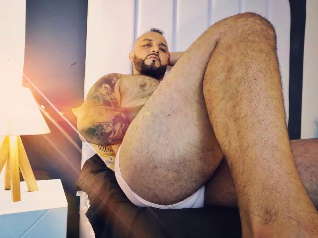 BardoCkroy69, muscular and hairy, sits with legs up, wearing white underwear, tattooed arm, and beard, looking at camera, sunlight on left, lamp on white nightstand.