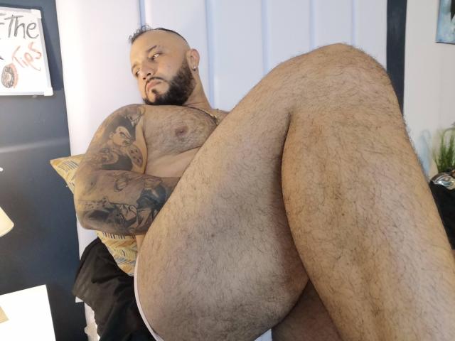 BardoCkroy69, a hairy, tattooed, bearded man with a bald head, is nude, sitting with legs up, showing his hairy ass and chest tattoos, looking at the camera.