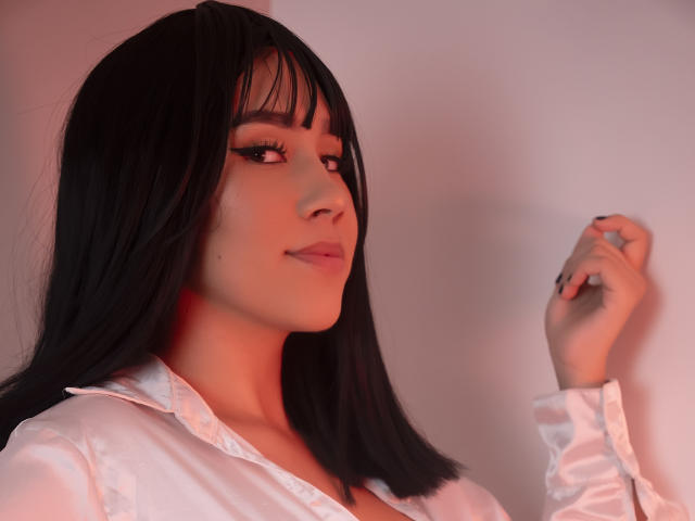 CelesteRusso has black hair, wearing a white shirt, with a slight smile, and raised hand, showcasing long, dark eyelashes and pink lipstick, in a soft, red light.